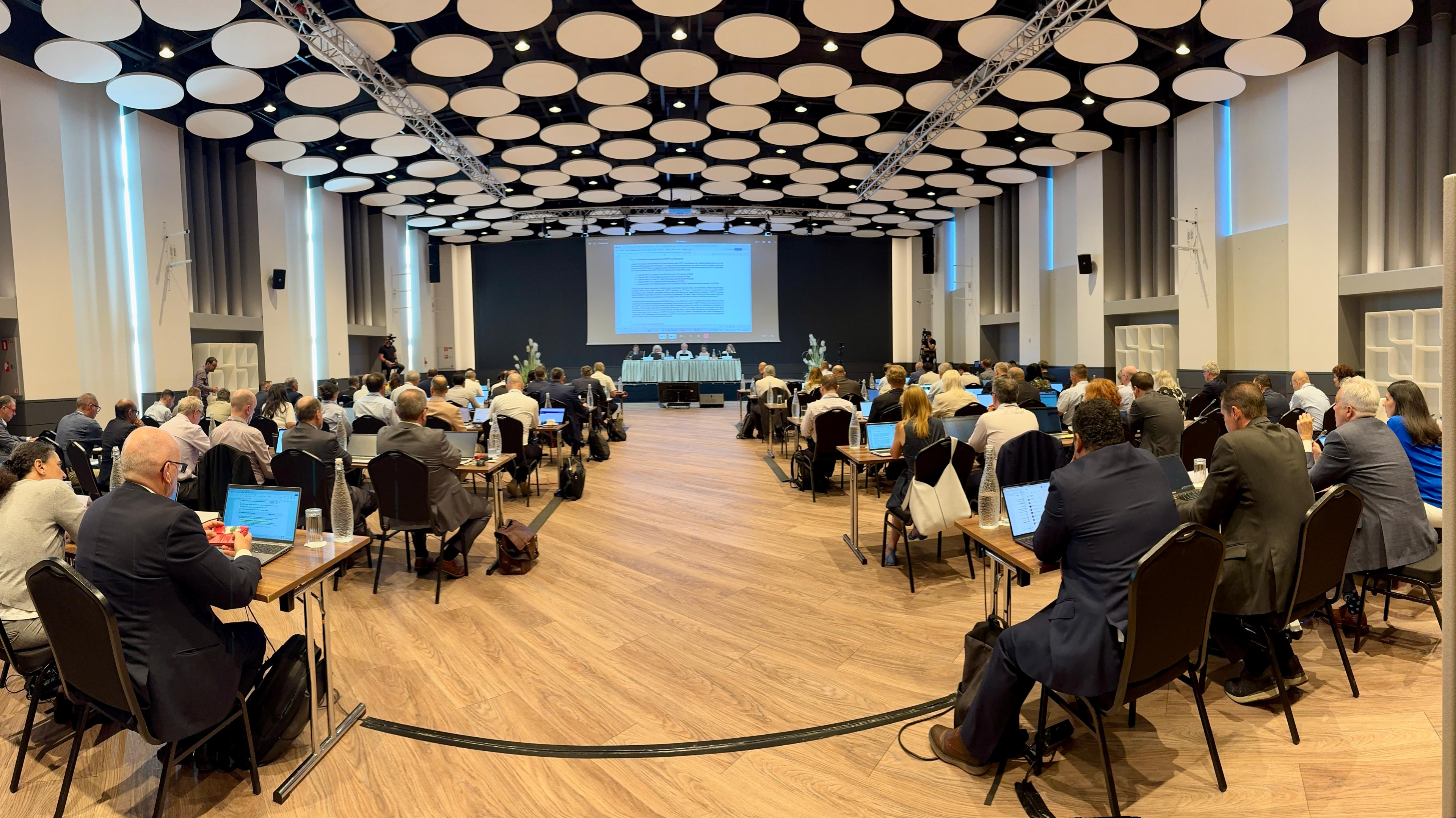 Konferenču zāle ar dalībniekiem pie galdiem, kuri klausās prezentāciju un diskusiju uz skatuves