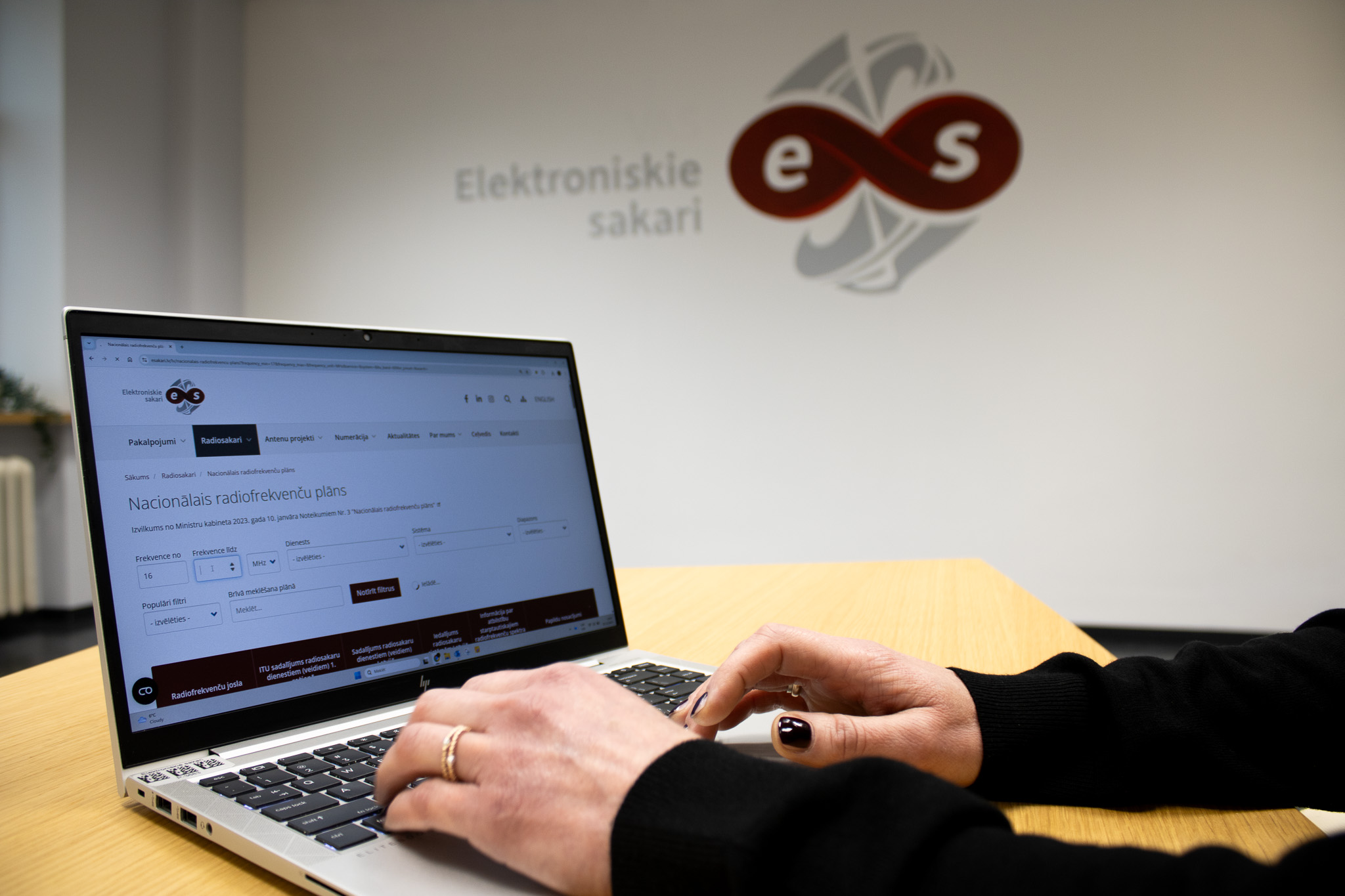 Persona izmanto datoru ar atvērtu Nacionālā radiofrekvenču plāna digitālo pārlūku, fonā redzams SIA Elektroniskie sakari logo