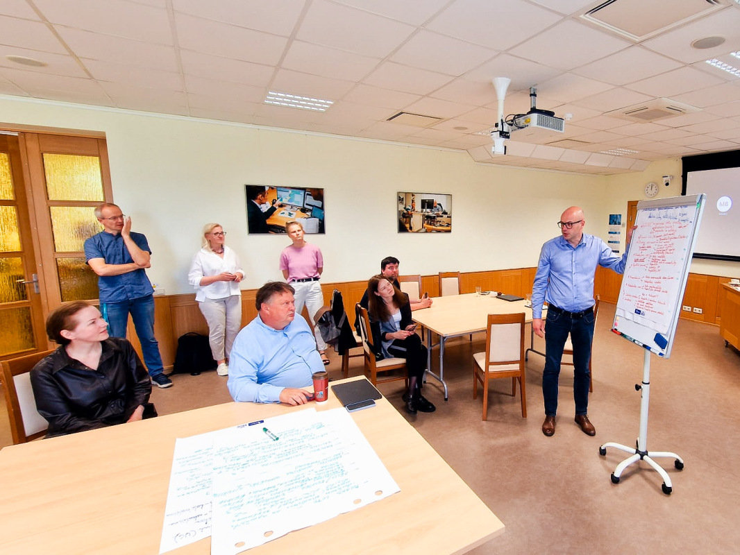 Attēlā redzama darba grupas sanāksme konferenču telpā. Telpā ir vairāki cilvēki – daži sēž, bet citi stāv. Vīrietis attēla labajā pusē pie baltās tāfeles vada prezentē idejas.