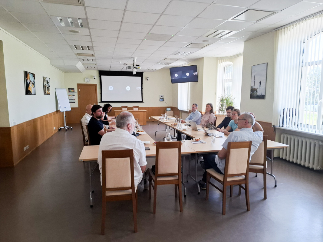Attēlā redzama sanāksme gaišā konferenču telpā, kur vairāki cilvēki sēž ap garu galdu, iesaistoties diskusijā. Uz galda redzami klēpjdatori, piezīmju bloknoti, ūdens glāzes un nelielas uzkodas. Fonā pie sienas atrodas divi ekrāni, kuros redzams tiešsaistes sanāksmes interfeiss ar pieslēgušos dalībnieku ikonām. Telpā ir koka apdare un sienas rotā fotogrāfijas, bet pie logiem ieplūst dabīgā gaisma.