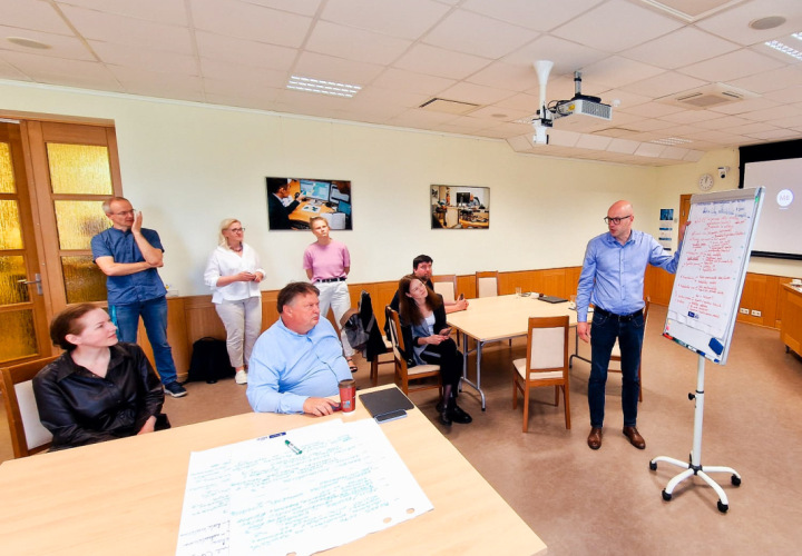 Attēlā redzama darba grupas sanāksme konferenču telpā. Telpā ir vairāki cilvēki – daži sēž, bet citi stāv. Vīrietis attēla labajā pusē pie baltās tāfeles vada prezentē idejas. 