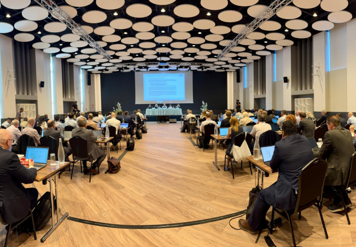 Konferenču zāle ar dalībniekiem pie galdiem, kuri klausās prezentāciju un diskusiju uz skatuves.