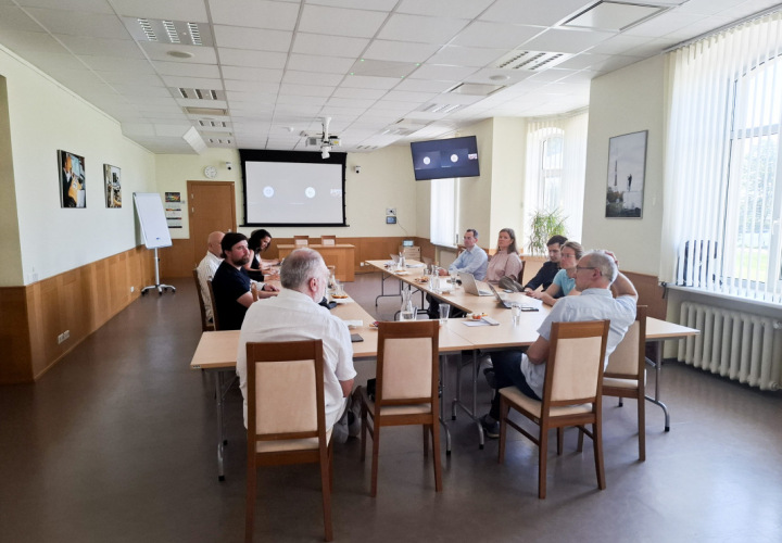 Attēlā redzama sanāksme gaišā konferenču telpā, kur vairāki cilvēki sēž ap garu galdu, iesaistoties diskusijā. Uz galda redzami klēpjdatori, piezīmju bloknoti, ūdens glāzes un nelielas uzkodas. Fonā pie sienas atrodas divi ekrāni, kuros redzams tiešsaistes sanāksmes interfeiss ar pieslēgušos dalībnieku ikonām. Telpā ir koka apdare un sienas rotā fotogrāfijas, bet pie logiem ieplūst dabīgā gaisma.