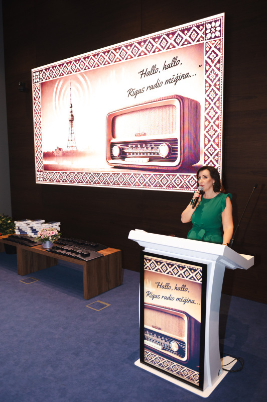 Sieviete zaļā kleitā runā pie tribīnes ar mikrofonu, fonā liels attēls ar radio un uzrakstu “Hallo, hallo, Rīgas radio mēģina…